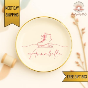 May include: A cream-colored decorative plate with a gold rim. The plate features a pink illustration of an ice skate and the name "Annabelle" in a cursive font. The plate is a decorative item.