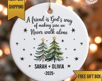 May include: A white ceramic Christmas ornament with a gold string for hanging. The ornament features a message, two Christmas trees, and the names SARAH + OLIVIA, with the year 2025. The text reads "A friend is God's way of making sure we Never walk alone."