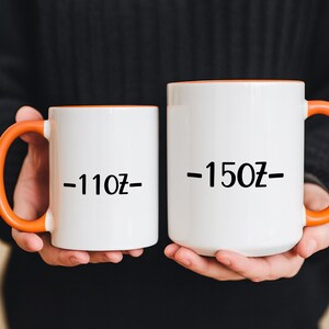 Two white mugs with orange handles are being held by a person wearing a black sweater.