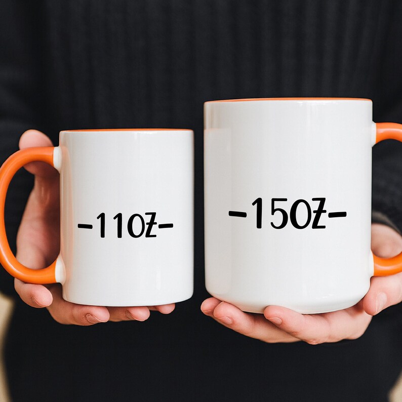 Two white mugs with orange handles are being held by a person wearing a black sweater.