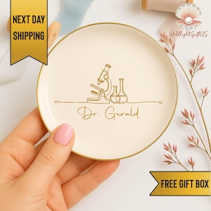 May include: A round, off-white ceramic trinket dish with a gold rim. The dish features a gold line drawing of a microscope and beakers, with the name "Dr. Gerald" below. The dish is held by a person.