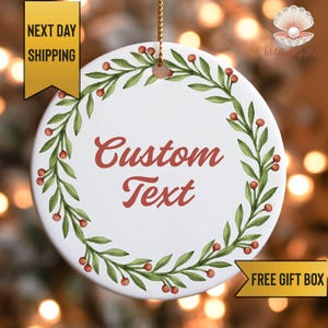May include: White ceramic ornament with a green and red botanical wreath design. The ornament has the words "Custom Text" in red script. A gold ribbon is attached for hanging. The words "Next Day Shipping" and "Free Gift Box" are also visible.