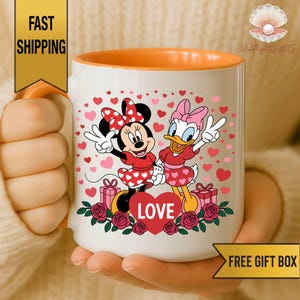A person is holding a white mug with an illustration of Minnie Mouse and Donald Duck surrounded by hearts and flowers.
