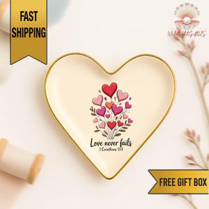 May include: Heart-shaped trinket dish with a gold rim, featuring a bouquet of red, pink, and coral hearts. The dish has the text "Love never fails" and "1 Corinthians 13:8".
