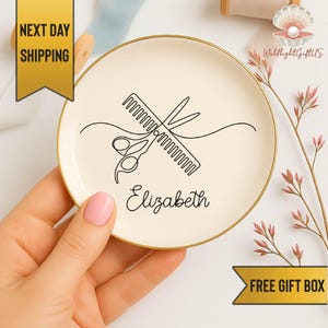 May include: A round, off-white ceramic trinket dish with a gold rim. The dish features a black line drawing of scissors and a comb, with the name "Elizabeth" below. The dish is held by a person with pink nail polish.