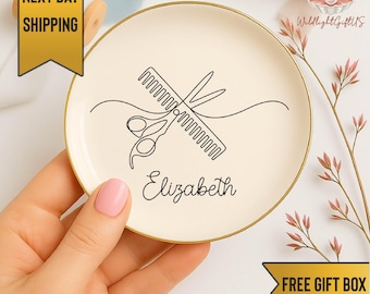 May include: A round, off-white ceramic trinket dish with a gold rim. The dish features a black line drawing of scissors and a comb, with the name "Elizabeth" below. The dish is held by a person with pink nail polish.