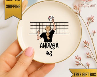 May include: A round, off-white decorative plate with a gold rim. The plate features a volleyball player illustration, a volleyball net, the name "ANDREA", and the number "3". The plate is held by a person with pink nail polish.