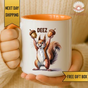 May include: A white ceramic mug with an orange interior and handle, featuring a graphic of a brown squirrel holding two acorns above its head with the text DEEZ printed in black letters above the squirrel's head on the front of the mug.