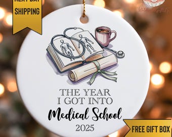 May include: White ceramic ornament with a medical school theme. It features an open book with anatomical drawings, a stethoscope, a coffee cup, and a rolled diploma tied with a green ribbon. Text reads "THE YEAR I GOT INTO Medical School 2025."