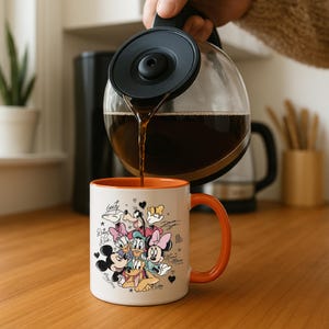 A hand is pouring coffee from a coffee maker into a white mug with an illustration of Disney characters on it.