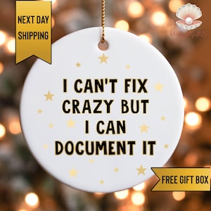 May include: A white ceramic ornament with the text "I CAN'T FIX CRAZY BUT I CAN DOCUMENT IT" in black. Gold stars are scattered around the text. The ornament has a gold ribbon for hanging. A gold banner reads "NEXT DAY SHIPPING".