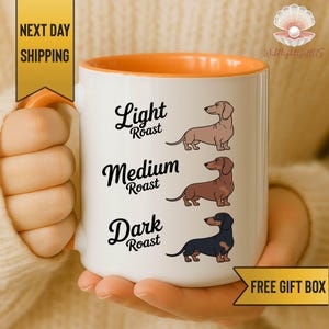 May include: White ceramic coffee mug with an orange interior and handle. The mug features illustrations of dachshunds and the words "Light Roast," "Medium Roast," and "Dark Roast." The mug is held by a person.