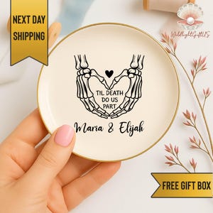 May include: A circular, cream-coloured ceramic dish with a gold edge. It displays a black graphic of skeleton hands forming a heart, with the words "TIL DEATH DO US PART" and the names "Maria & Elijah".
