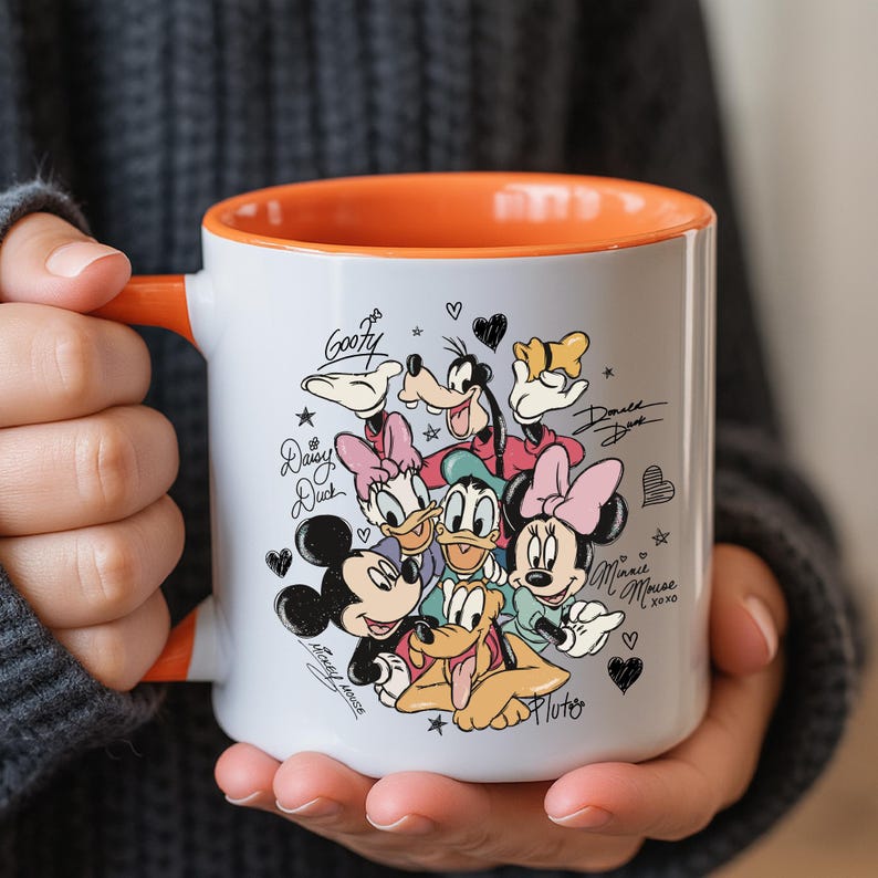 A person is holding a white mug with an illustration of Disney characters, including Goofy, Minnie Mouse, Mickey Mouse, Donald Duck, and Pluto.
