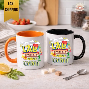 May include: Two white ceramic mugs with colorful designs. One mug has an orange handle and interior, the other has a black handle and interior. Both mugs feature the text "LAB STORY 2-26 EMMA" in a playful, colorful font, with science-themed graphics.