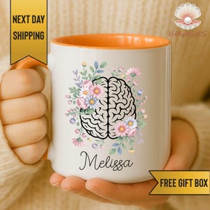 May include: A white ceramic mug with an orange interior and handle. The front features a black line-art illustration of a human brain surrounded by colorful pink, blue, and white flowers and green leaves. The name Melissa is printed in black script.