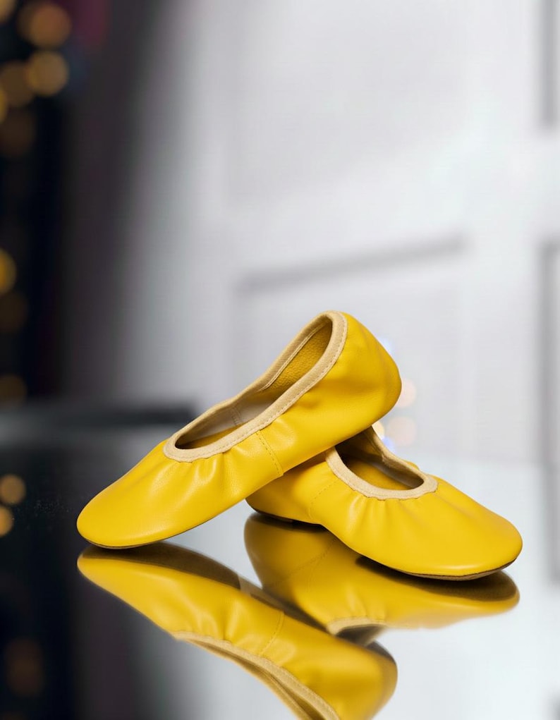 May include: A pair of bright yellow ballet flats with a rounded toe and a cream-colored trim. The shoes are made of a soft material and are reflected in the shiny surface below.