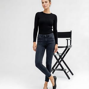 May include: A woman wearing a black ribbed long-sleeve top and dark wash skinny jeans. She is also wearing black flats. A black director's chair is behind her.