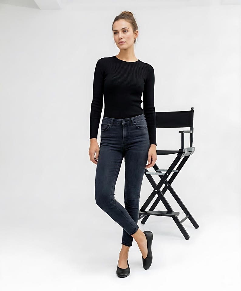 May include: A woman wearing a black ribbed long-sleeve top and dark wash skinny jeans. She is also wearing black flats. A black director's chair is behind her.