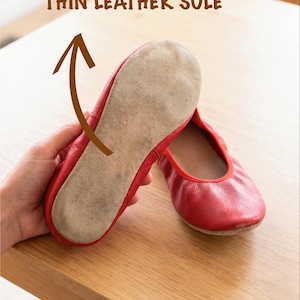 May include: Red ballet flats with thin leather soles. One shoe is held up to show the sole, with the text "THIN LEATHER SOLE" above. The other shoe is on a wooden surface.