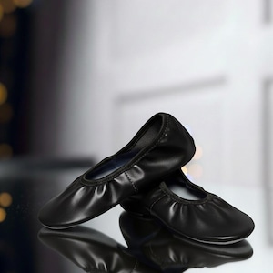 May include: A pair of black ballet flats with a rounded toe and a gathered vamp. The shoes are made of a smooth, shiny material and are reflected in the glossy surface below. The background is blurred, with hints of light.