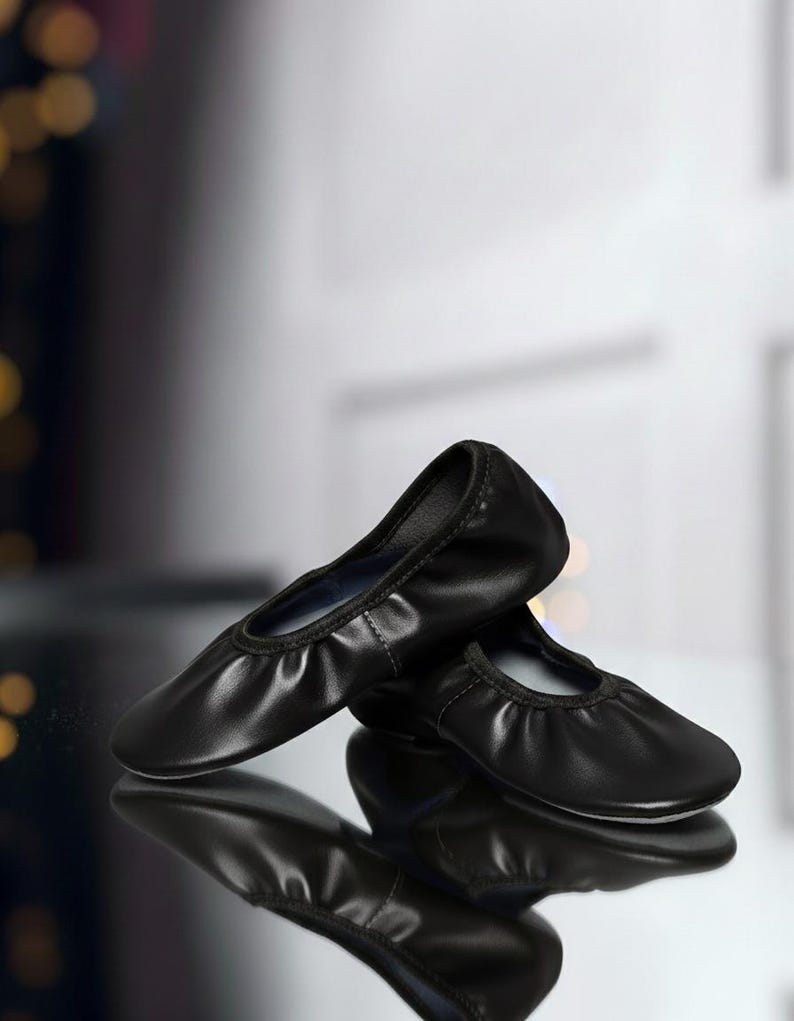 May include: A pair of black ballet flats with a rounded toe and a gathered vamp. The shoes are made of a smooth, shiny material and are reflected in the glossy surface below. The background is blurred, with hints of light.