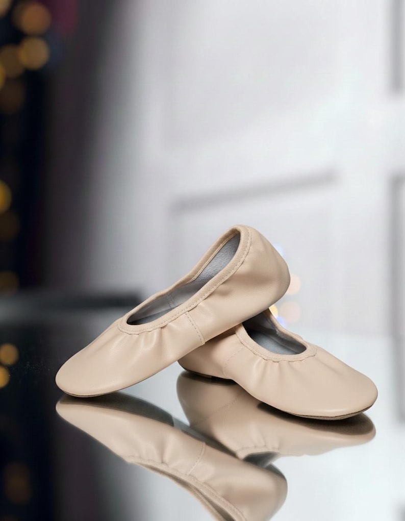 May include: A pair of beige ballet flats. The shoes are made of a soft material and have a rounded toe. The soles are thin and flexible. The shoes are designed for dance or casual wear.