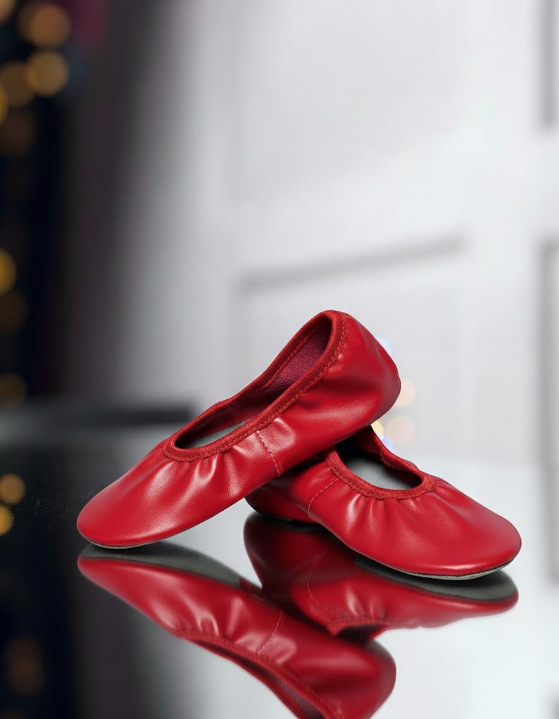 May include: A pair of red ballet flats. The shoes are made of a smooth material and have a rounded toe. The flats are reflected on a shiny surface. The background is blurred, with hints of light and shadow.