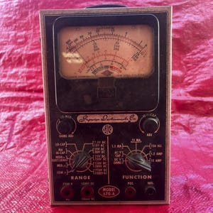 Peut inclure: Un multimètre vintage Superior Instruments Co. Model 670-A doté d'un grand cadran analogique avec plusieurs échelles, deux boutons de commande rotatifs noirs et divers ports d'entrée étiquetés. L'appareil possède une façade noire avec des inscriptions blanches.