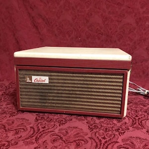 Puede incluir: Radio Capitol vintage con tapa color crema y marco rojo. La parte delantera presenta una cubierta de altavoz tejida en oro y marrón. La radio tiene una pequeña etiqueta que dice "Capitol". Un cable de alimentación se extiende desde el lateral.