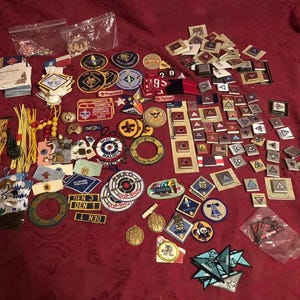 Op de afbeelding: Een verzameling van padvindersbadges, patches en spelden in verschillende vormen en maten. De items hebben een reeks kleuren en ontwerpen, met tekst en symbolen. Sommige items zitten in kleine plastic zakken.