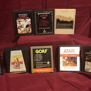 Può includere: Una collezione di otto cartucce rettangolari per videogiochi Atari disposte su due file. Le cartucce presentano varie etichette tra cui Combat, Gorf, Popeye, Shark Attack e Super Breakout, con involucri in plastica nera, bianca e color sabbia.