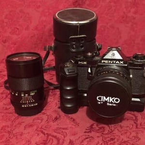 Puede incluir: Una cámara Pentax negra con una lente y una tapa de lente negra que dice "CIMKO MY Series". Una lente negra está a la izquierda, y una funda de cuero negra está detrás de la cámara. La cámara está sobre una superficie roja estampada.