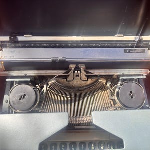Puede incluir: Máquina de escribir vintage abierta, con carro plateado, teclas negras y marco color crema. El texto "PAGE GAGE" es visible en el carro. La máquina de escribir está en su estuche de transporte abierto.