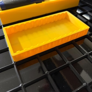 May include: A bright yellow tool organizer with a rectangular shape and a series of vertical ridges along the sides. The organizer is sitting on a black, grid-patterned surface. Several chisels with blue and black handles are visible in a similar yellow container.