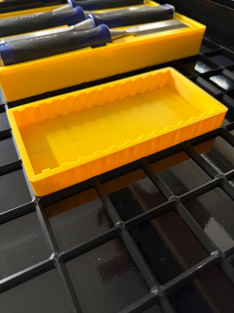 May include: A bright yellow tool organizer with a rectangular shape and a series of vertical ridges along the sides. The organizer is sitting on a black, grid-patterned surface. Several chisels with blue and black handles are visible in a similar yellow container.