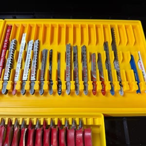 May include: A yellow toolbox with a variety of jigsaw blades. The blades are in a yellow tray with individual slots. The blades are various sizes and have red, white, and silver accents. Some blades have text that reads "General Purpose" and "Reverse Cut Wood".