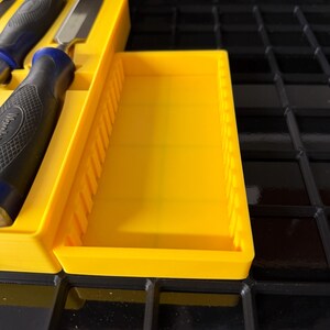 May include: A yellow plastic tool organizer with a rectangular shape and a series of vertical dividers. The organizer holds several chisels with black and blue handles. The chisels are made of metal and have a sharp, angled edge.