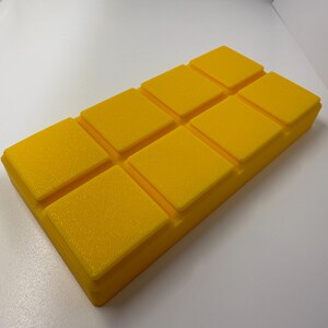 May include: A bright yellow, rectangular object with a textured surface. The object is divided into eight square sections, each with a slightly raised edge. The overall shape resembles a bar of soap or a block of wax.