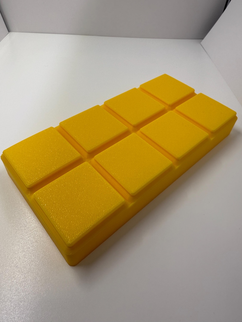 May include: A bright yellow, rectangular object with a textured surface. The object is divided into eight square sections, each with a slightly raised edge. The overall shape resembles a bar of soap or a block of wax.