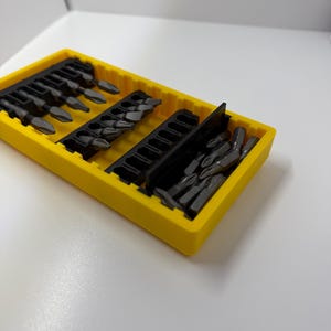 May include: A yellow plastic organizer filled with various dark gray screwdriver bits. The bits are arranged in rows within the organizer. The organizer is on a white surface.