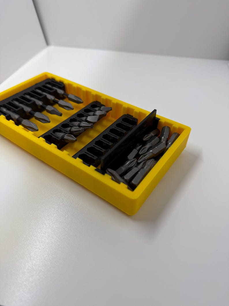 May include: A yellow plastic organizer filled with various dark gray screwdriver bits. The bits are arranged in rows within the organizer. The organizer is on a white surface.