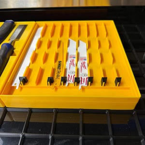 May include: A bright yellow plastic storage tray containing several metal reciprocating saw blades. The blades include one yellow DeWalt 6-inch blade and two white Milwaukee Sawzall metal-cutting blades, all organized in individual slots.