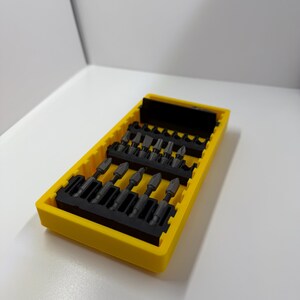 May include: A yellow plastic case containing a set of black screwdriver bits. The bits are arranged in rows within the case. The case is rectangular and has a slightly textured surface.