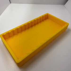 May include: A bright yellow rectangular tray with a textured interior and a series of vertical ridges along one long edge. The tray is made of a solid material and sits on a white surface.