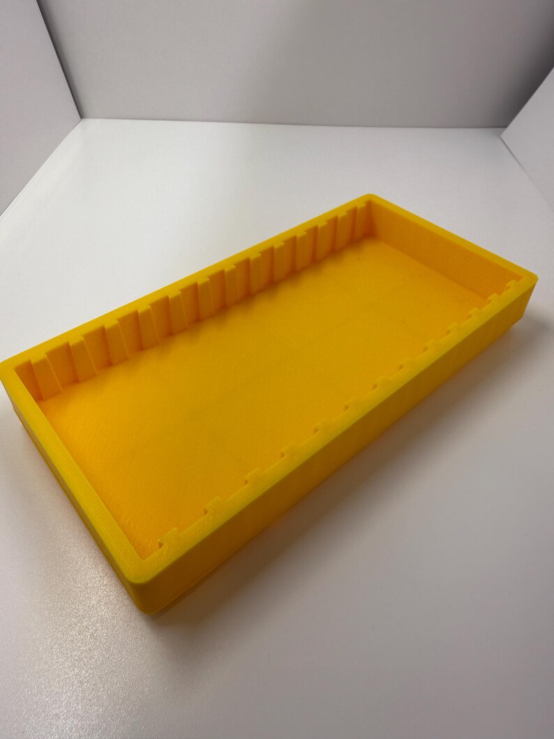 May include: A bright yellow rectangular tray with a textured interior and a series of vertical ridges along one long edge. The tray is made of a solid material and sits on a white surface.