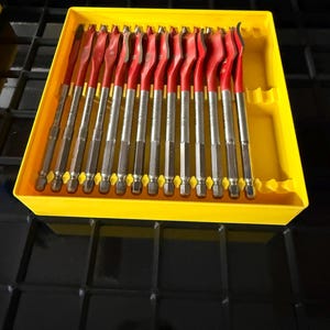 May include: A set of thirteen spade drill bits with red-painted shanks and silver-colored tips, arranged in a bright yellow plastic storage tray. Each bit features a hexagonal shank and a flat, wide cutting blade designed for drilling holes in wood.