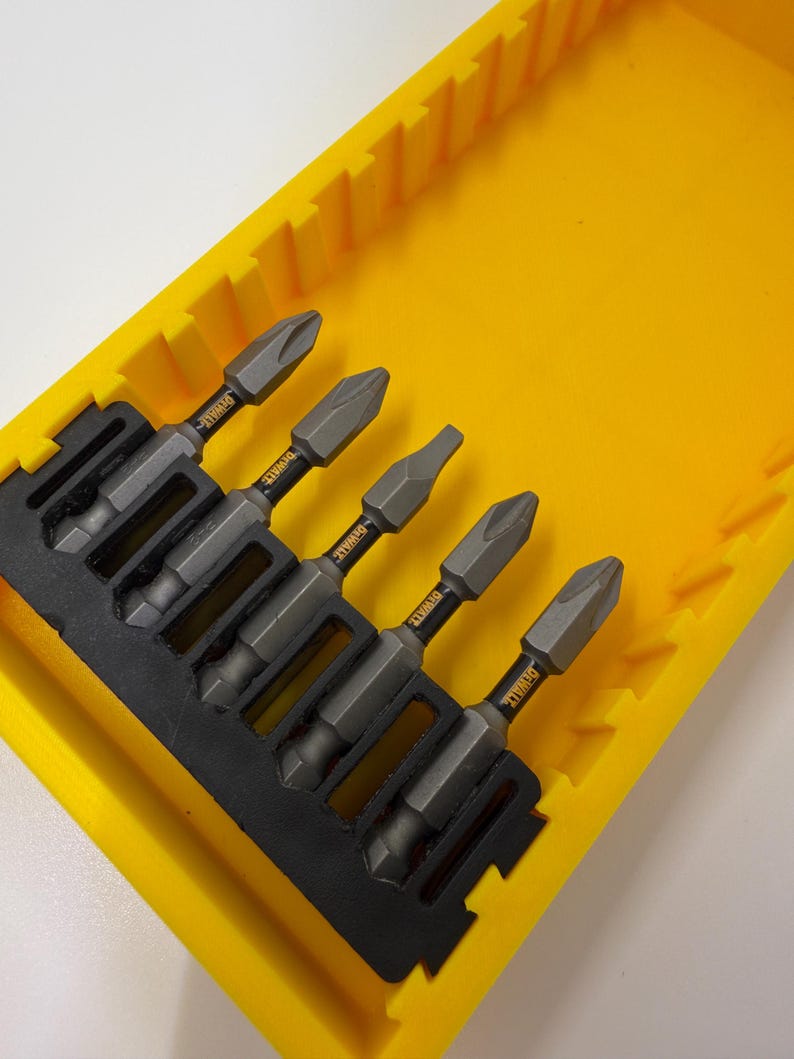 May include: A set of five gray and black screwdriver bits with the brand name "DEWALT" printed on them, neatly arranged in a bright yellow plastic case. The case has a black insert to hold the bits securely.