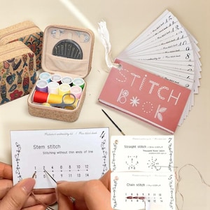 Peut inclure: Un kit de couture avec une variété de fils colorés, des aiguilles et des ciseaux. Le kit comprend un petit étui ouvert avec un intérieur beige et une collection de bobines de fil. Un "Stitch Book" et des cartes de guide de points sont également présents.