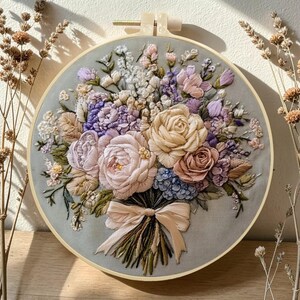 Puede incluir: Un bordado detallado de un ramo floral en un aro redondo. El ramo presenta rosas, hortensias y otras flores en tonos de rosa, morado y crema, atado con una cinta. El fondo es de tela gris claro.